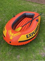 Orange and yellow Explorer 300 inflatable dinghy with one paddle lying on the grass. Shows overall condition and colors.