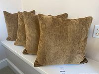 Four gold-colored cushions with herringbone pattern arranged in a row on a white shelf against a wall.