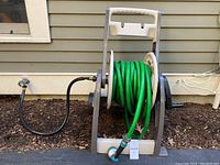 Green garden hose wound onto a plastic Hose Mobile reel with crank handle, set next to house exterior wall