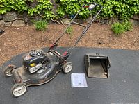 Side and front view of the lawn mower showing overall shape, wheels, grass catcher, and instruction manual on ground.