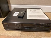 Front view of Onkyo DX-C3306 6-disc CD changer showing power button, display screen, disc selection and transport controls, with remote and instruction manual on top.