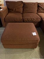 Front view of the brown upholstered ottoman matching the sectional couch, showing its soft cushioned top and fabric texture.