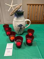Lot consisting of a Stone Soldier pottery jug decorated with sea-themed ornaments and six red glass votive holders containing candles, displayed on a green tablecloth.