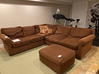 Brown fabric sectional sofa with chaise shape showing all three sections together with an ottoman in front. Located in basement or similar room.