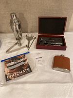 Full set of barware items arranged on white surface including cocktail shaker, jigger, tongs, spoon; wooden box with wine tools logo; brown leather flask; Chitchats drink markers in plastic bag.