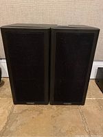 Front view of both black Mission speakers with fabric grilles and Mission logos at bottom front.