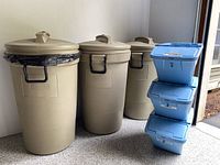 Three beige Rubbermaid trash barrels on wheels lined up against a wall next to three blue stackable recycling containers with lids stacked vertically.