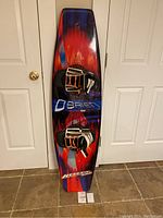 Top view of OBrien Heretic 140 wakeboard showing attached black and white Blindside boots with orange laces on the red and blue patterned board.