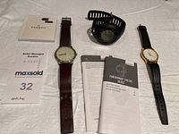 Photo showing the Skagen Time wristwatch with black leather strap, brown leather strap analog watch, Garmin watch with manuals and papers configured on white cloth surface.