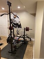 Full setup on black rubber mat, showing bench, lat tower, loaded barbell, weight plates underneath