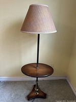 Full view of the antique one-piece table lamp with beige fabric lampshade on top, showing the dark metal rod and round wooden table surface, tripod base with metal details.