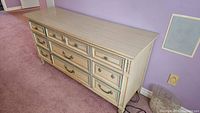 Front angled view of dresser showing nine drawers, cream wood, vintage style with decorative handles and trim
