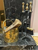 Photo showing brass fireplace tool set including brass hammered log holder with handle, broom brush, and three brass-handled tools (shovel, poker, tongs) on black metal stand.