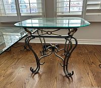 End table shown from a side angle, highlighting the glass top and metal stand with intricate scrollwork and circular detail in the center.
