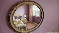Photo of oval hanging mirror with decorative frame, reflecting bedroom scene with purple walls, nightstand, lamp and bed.