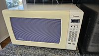 Front view of Panasonic microwave showing beige exterior, digital control panel and door with mesh window.