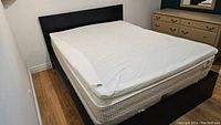 Black IKEA queen bed frame with beige mattress and white fitted sheet, assembled in a small bedroom corner.