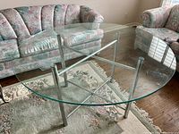 Round glass table set with clear glass top and silver metal base, placed on floral rug between pink and green upholstered sofas