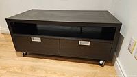 Front view of black TV stand showing two drawers, open shelf, and caster wheels, positioned against a white wall on a wooden floor.