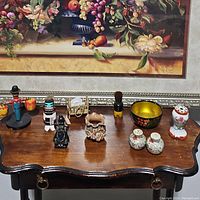 Top-down view of assorted figurines, dishes, salt and pepper shakers arranged on a dark wooden curved edge surface against a floral framed picture background.