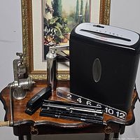 Photo showing a mix of office supplies including staplers, hole punch, numbering stamp device, and a black and silver paper shredder on a wooden small table with a framed picture behind.