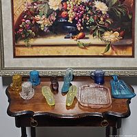 Overview of ten vintage glass pieces arranged on small wooden table under a framed floral painting.