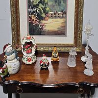 Table displaying seven Christmas decorative pieces including ceramic snowman, Santa shaker, reindeer, ceramic house ornament, glass lamp post, and two glass angel figurines.