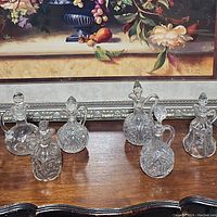 Photo showing six clear glass cruets with various cut and pressed glass patterns and stoppers on a wooden table in front of a framed floral painting.