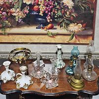 Full lot of mixed oil lamps and candle holders arranged on a wooden table with a floral painting background. Includes various glass and metal lamps and candle holders.