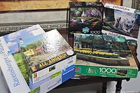 View of seven assorted 1000-piece puzzle boxes stacked together featuring wildlife, lighthouse, train and nature scenes.