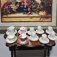 Ten assorted tea cups and saucers placed on a wooden table in front of a floral painting, showing variety of floral and gold designs.