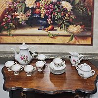 Wide view of the tea set on a wooden surface, showing teapot, six cups, six saucers, creamer, and sugar bowl with floral rose design