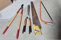 Photo showing five tools including loppers, pruning shears, garden shears, handsaw, and bow saw on concrete floor.