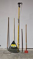 Full view of five assorted garden and hand tools against a white wall including a small metal hand rake, black plastic leaf rake, yellow post hole digger, metal pick, and red square head tool.