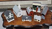 Seven different building pieces for train accessories include a church, house, barn, silo, and assorted buildings in various colors and shapes laid on a wooden table.