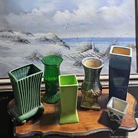 Six assorted vases displayed on a wooden surface in front of a coastal landscape painting, showing variety in shape, color, and material.