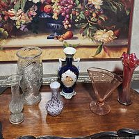 Photo showing all six assorted vases on a wooden surface in front of a framed painting.