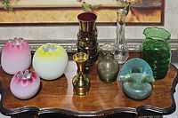 Overview of all nine vases showing variety in color, material, and size on wooden table against framed artwork background.