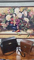 Two carrying cases, one brown leather and one black textured, placed on a wooden table in front of a floral painting.