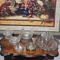 Clear pressed glass dishes on wooden table with floral painting background, showing assortment of 7 glass dish items including the hen-shaped bowl, covered butter dish, round scalloped dish, floral patterned bowls, small round dish, and glass cake dome with base.