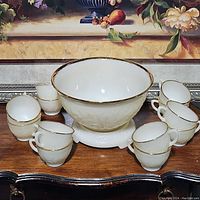 Complete vintage punch bowl set consisting of one large embossed white ceramic bowl with 22k gold trim and eight matching cups arranged around it.