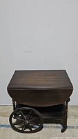 Full front view of vintage wooden tea cart with drop-leaf sides folded down, showing large wooden wheels and lower shelf.