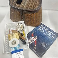 Photo showing antique wicker leather fishing creel basket, vintage fishing reels, Gerber Clutch Shortcut multi-tool, fishing line spool, accessories and fly fishing book.