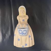 Front view of the ceramic girl figurine showing bonnet, dress, and basket details with blue flower and bow motifs.