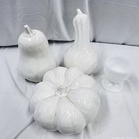Three white ceramic decorative vegetable sculptures and one white milk glass cup arranged on a cloth background. Shows pumpkin, pear, gourd sculptures and cup.
