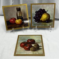 Photo of three vintage fruit motif lithograph prints by Henk Bos displayed standing on clear easels. Each print has a golden border and depicts different fruit arrangements in still life style.
