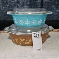 Two stacked Pyrex casseroles, turquoise with white snowflakes on top and brown with gold Early American patterns below, both with glass lids.