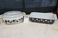 Pair of Pyrex casseroles: oval Black Tulip with glass lid and metal holder, and rectangular black casserole with white snowflake pattern.