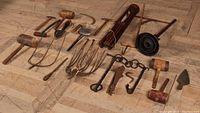 Overview of the collection spread out on wooden floor showing variety of primitive farm implements including mallets, sickle, shovel, and irons.
