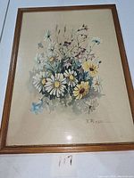 Framed B. Riasni botanical flower print showing daisies, small wildflowers, and greenery on beige background in a brown wooden frame.